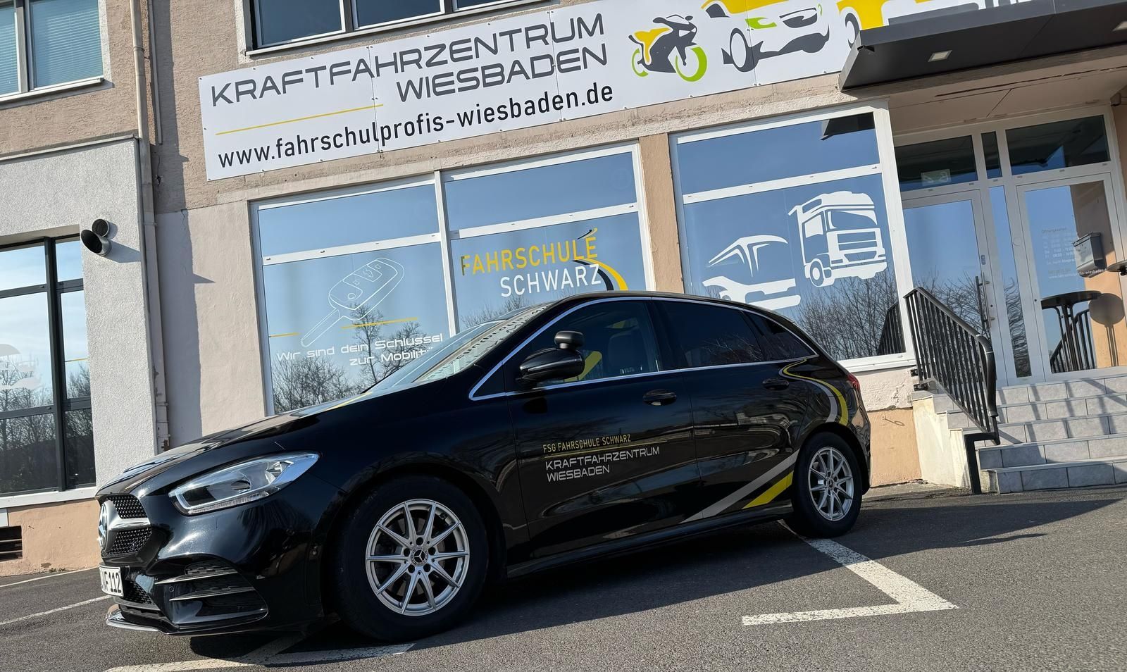 Ein schwarzes Auto, das vor dem Kraftfahrzentrum Wiesbaden geparkt ist und das Branding und die Außenbeschilderung der Fahrschule zeigt.